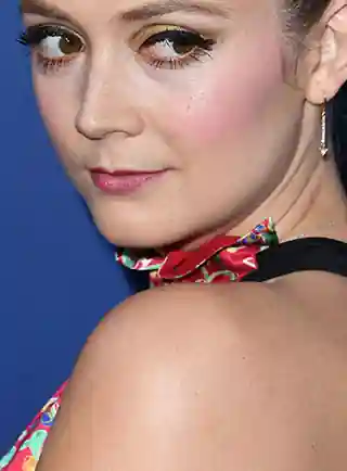 Billie Lourd turns her head. She is seen in front of a purple wall. Billie wears a red, green and black floral print gown. She is seen with her hair tied up in a bun. Billie wears a dark pink lipstick, black mascara and a pair of dangling earrings.