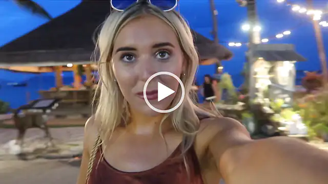 Em Sheldon holds a camera at arms length as she vlogs on holiday. She wears a pair of white frame sunglasses on her head with her hair loose. She appears in a copper satin dress against a deep blue sky at dusk.