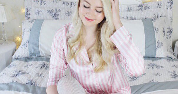 Meg Says wearing a pink and white stripe satin pyjama top on a grey, blue and white bed. She is touching her hair that is loose and slightly wavey.