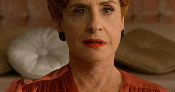 Patti LuPone appears on a luxurious cream and pink velvet bed. She wears a dark orange satin dress and large black earrings with her hair pinned upwards.