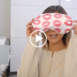 Naomi Victoria holds a white and red kiss pattern eye mask in front of her face. She wears a thin jumper in a white and cream room.