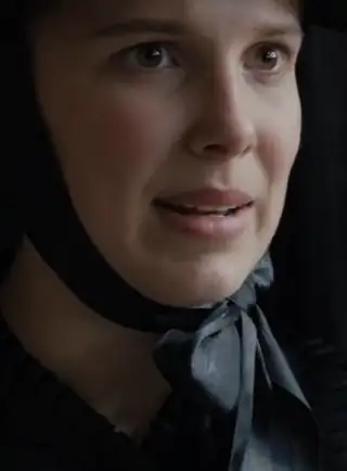 Millie Bobby Brown wears a black bonnet tied at her chin with a large black ribbon. She wears soft foundation with a blush pink lipstick.