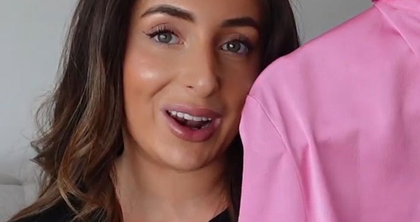 Hazel Maria Wood holds up a bright pink satin shirt. Her hair is loose and curled and she appears in front of a grey wall wearing a black top.