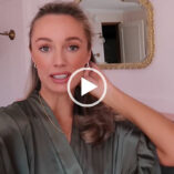 Josie Irons is seen in a dark green satin dressing gown. She reaches back and touches her loose hair. Josie wears small hoop earrings and a gold necklace in a pale pink room.