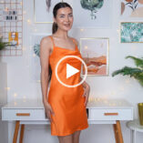 Roxxsaurus appears in her white and light wood filming area at home. She has her hair tied back in a ponytail. Roxi stands and looks into the camera with her hands by her sides. She wears a bright orange dress from Primark.