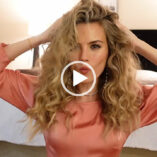 Niomi Smart runs both her hands through her curled and volumised hair. She sits in front of a large white and grey bed in a room that has cream wardrobes and soft lighting. Niomi wears a long sleeve peach gown in London as she gets ready for an event.