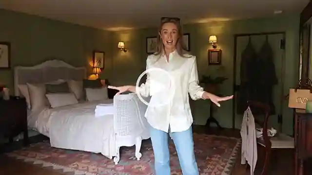 Josie Fear stands in a green and dark wood bedroom at her countryside home with her hands outstretched. She wears a long sleeve shirt with a pair of light blue jeans. Josie wears her long blonde hair straightened and loose with a pair of black sunglasses on her head.