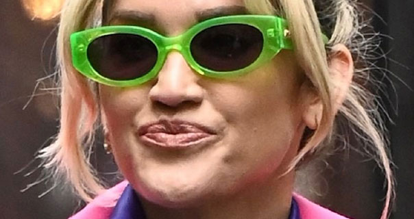 Ashley Roberts appears in a pair of neon green large frame sunglasses. She wears a pink coat with a purple satin top.