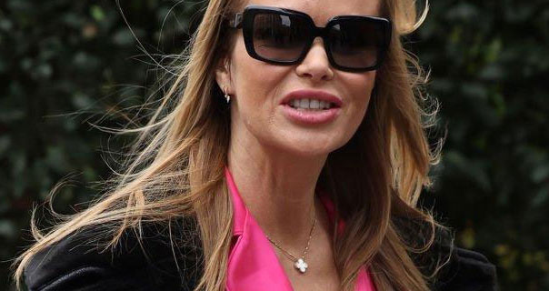 Amanda Holden appears in a thick black winter coat and pink satin. She wears a pair of sunglasses against a dark green leafy background.