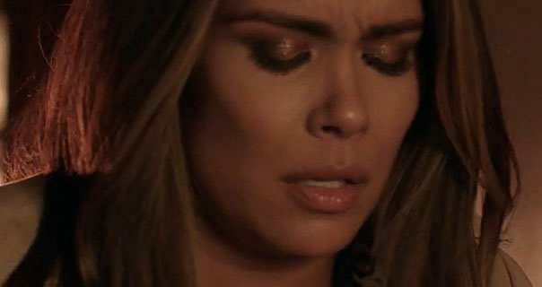 Daniella Alonso closes her eyes showing pain in an episode of Dynasty. She wears a trench coat and leans against a wall in orange lighting in a hallway.