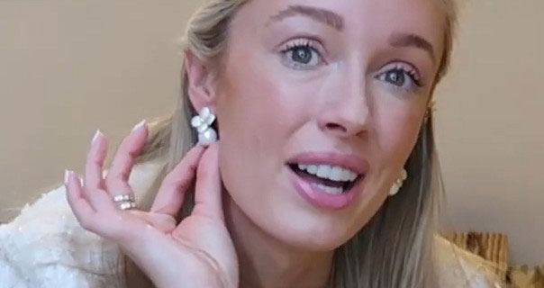 Josie Irons touches a pearl earring she is wearing. She appears in a fluffy cream jacket against a light brown wall.