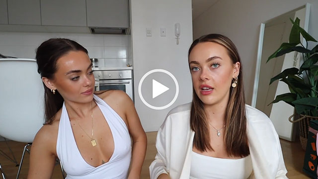 Ashley and Olivia Mescia sit on the floor in an open plan living room. Ashley looks at Olivia who is wearing a light yellow dressing gown over a white top. Ashley wears a white halter neck plunge top and her hair is tied back.