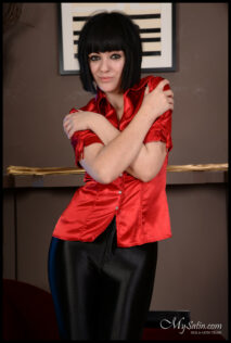 Scarlet Smith stands with her hands crossed touching her shoulders. She wears a red satin short sleeve blouse and a pair of shiny black trousers.