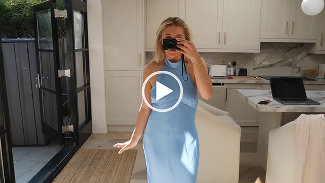 Em Sheldon stands in her kitchen with her patio doors open at the side. She records herself through a full length mirror. Em wears a baby blue satin halter neck dress. Her her is loose and back and she holds her vlogging camera in front of her face.