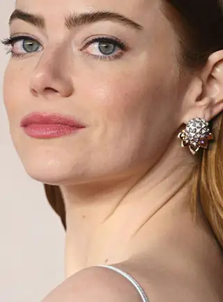 Emma Stone turns her head towards the camera. She wears her hair back and behind her ears. She appears in large silver earrings against a neutral background.