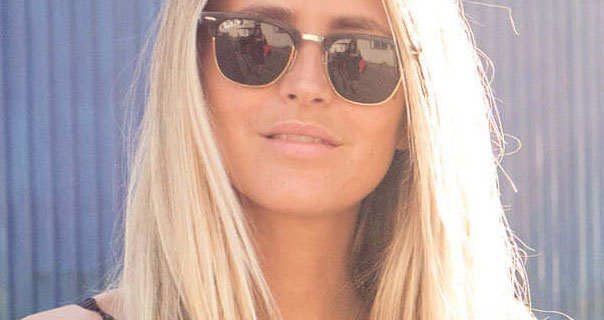 Janni Delér wears a pair of black and gold sunglasses outside in front of a blue grey corrugated metal wall. Her hair frames her face and is light by bright sunshine.