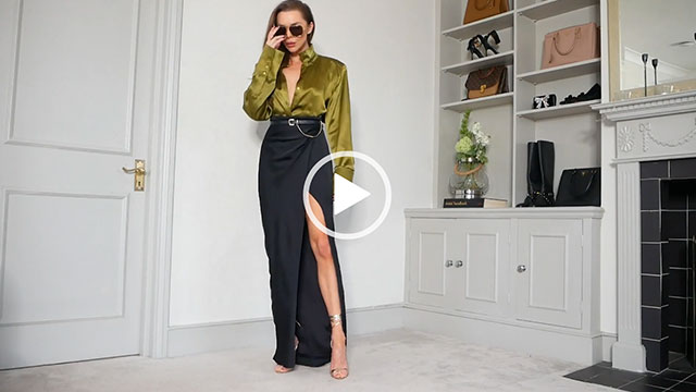 Laura Blair adjusts her sunglasses. She wears an unbuttoned olive green satin long sleeve shirt and a long high slit black skirt. She appears in her grey and white living room near a black tile fireplace.