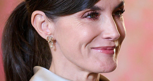 Queen Letizia Of Spain smiles slightly. She wears light grey satin with a pair of dangling earrings against a pale red background.
