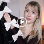 Meg Says slides her hand through a black velvet and white satin ribbon dress. She wears a black long sleeve top with her blonde hair loose. Meg appears against a white bedroom backdrop with a dark wooden chest of drawers and a small lit candle behind her.