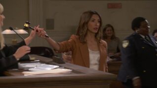 India de Beaufort hands a gavel to Melissa Rauch in a courtroom with the jury behind her. She wears a blazer and a white satin camisole top.