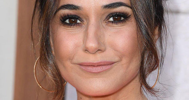 Emmanuelle Chriqui smiles with her mouth closed. She wears a pale pink lipstick with large gold hoop earrings against a white and pale brown background. Her hair is tied up with curled strands framing both sides of her face.