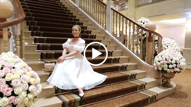 Freddy Cousin-Brown sits on a grand brown and cream staircase at Disneyland Paris. She wears a white satin and silk puff sleeve gown with a leather back and satin shoes.