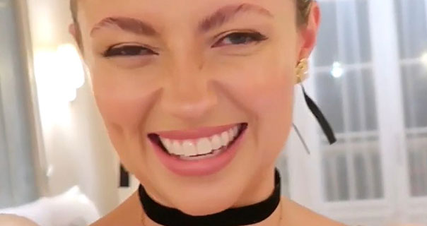 Freddy Cousin-Brown smiles into her camera as she vlogs. She wears her hair tied up in a Disneyland Paris hotel room. She is seen with a black ribbon necklace, black ribbon hair tie and gold earrings against a cream background with large white windows.
