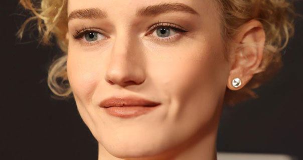 Julia Garner wears a large diamond earring with her curly hair short. She smiles a little against a black background with a small amount of pale gold in the lower corner.