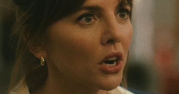 Ophelia Lovibond is seen with her hair up against a blurred red and light blue background. She wears a thick gold hoop earring. The collar of the cream satin shirt she is wearing can just be seen.