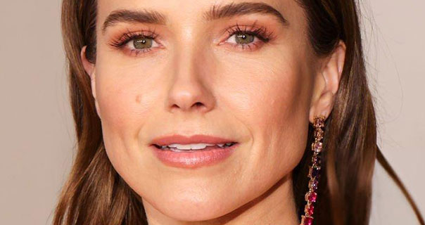 Sophia Bush looks straight ahead in front of a beige background. Her hair is loose and straight and is tucked behind her ears. She wears a pair of long dangling red jewel earrings.