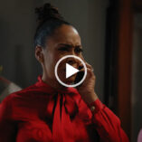 Simone Missick scowls as she talks into a mobile phone she is holding up to her ear. She wears her hair tied up in a bun. She is seen in a red satin top against a dark grey background.