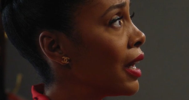 A side profile of Simone Missick in front of a dark grey background. Simone wears a red satin top, gold Chanel logo earrings and dark red lipstick.