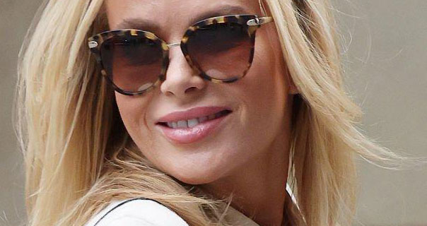 Amanda Holden turns her head. She appears against a taupe background with her hair down and blowing in the wind. Amanda wears a cream coat and large tortoiseshell sunglasses.