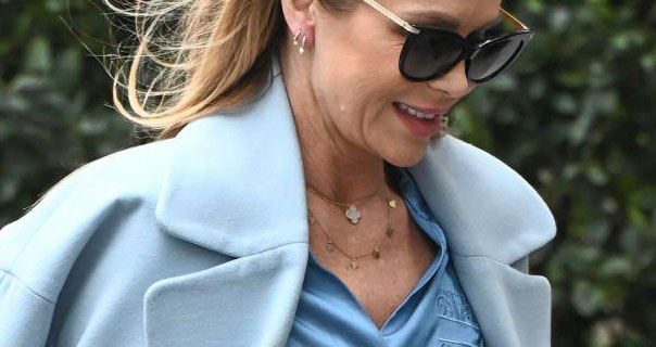 Amanda Holden wears a blue satin top with a pale blue trench coat. She pairs satin with sunglasses. Her hair is loose and blows in the wind as she walks by a green hedge.