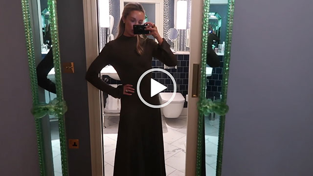 Josie Irons holds a camera up as she records in a London hotel room. She wears a dark green gown with her hair tied back and a hand on her hip. She is seen alongside a light purple and green wall with a blue tile and white marble bathroom behind her.