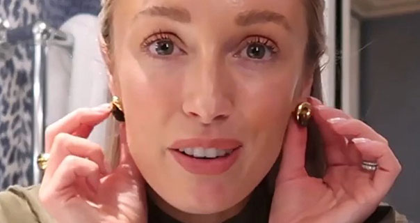 Josie Irons touches the gold earrings she is wearing. She appears in a dark green top in front of an animal print wall in a London hotel bathroom.