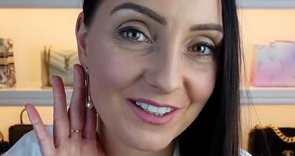 Steph Turton holds her hand up to frame the dangling HELAS pearl earring she wears. She appears in a white satin top in front of her handbag display.