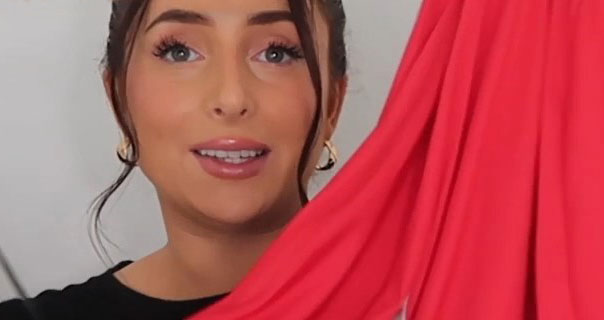 Hazel Maria Wood holds up a lightweight red top. She has her hair tied up and wears a pair of chunky gold hoop earrings against an dark white background.