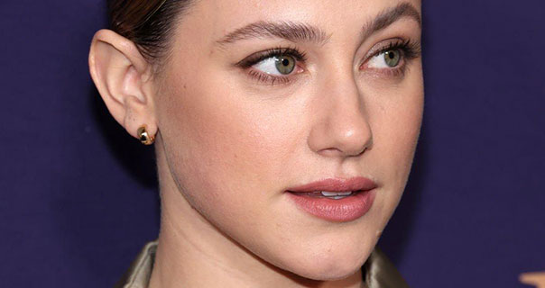 Lili Reinhart looks to the side. She appears in a dark gold satin shirt in front of a deep purple wall. She wears a pair of small chunky circle gold earrings with her hair tied back in a tight bun.