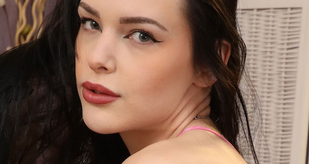 Antonia Jay looks over her shoulder into the camera. The strap of her pink satin gown is seen. She wears her black hair loose in front of a white and pale purple background.