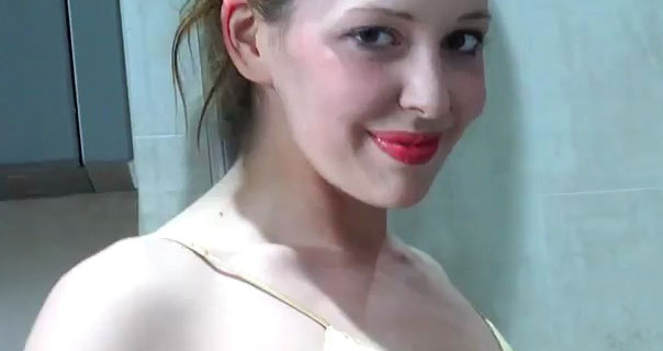 Alora Lux wears a gold satin nightie and red lipstick. She looks at the camera, appearing in a bathroom of white tiles and grey cabinets.
