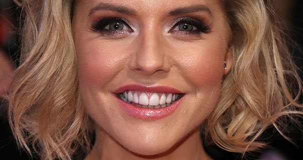 Stephanie Waring looks dead ahead with a wide smile. She is wearing pink lipstick and her blonde hair is loose and curled. She appears against a black background.