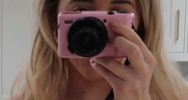 Em Sheldon holds her pink vlogging camera in front of her face. She has her hair loose and slightly curled as she films in the white marble kitchen in her London flat.