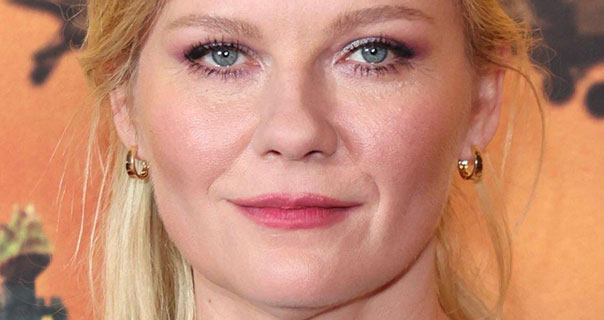Kirsten Dunst wears a pair of small gold hoop earrings with her hair tied back. She is seen with pink lipstick against an orange and black background.