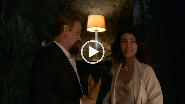 Jewel Staite turn to speak. She wears a cream jacket with a cream silk and lace camisole top. Jewel appears in a room in a cave with a small lamp and a chest behind her.
