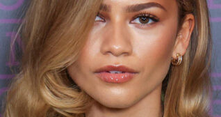 Zendaya is seen with her hair loose and curled in front of one eye. She wears glossy lipstick and a small gold hoop earring. Zendaya appears in front of a light and dark purple background.