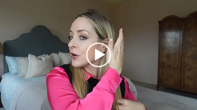 Fleur De Force runs her hands through her hair which is sleek and straight. She wears a pink satin shirt with a black sleeveless jumper. Fleur sits in one of the guest bedrooms in her home with an antique wardrobe behind her.