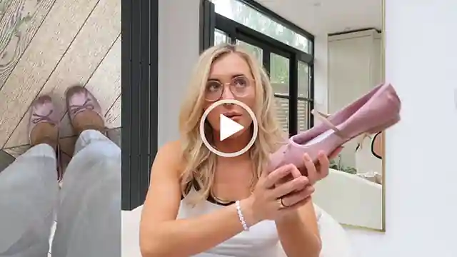 Em Sheldon glances upwards as she films in her kitchen. She holds a pair of pink ballet flats with her hair down and curled at the ends. Em wears a white top that has black trim, a pearl braclet and a single gold ring. She sits on a cream chair in front of a large mirror and the frame is split with a cutaway of her wearing the shoes.
