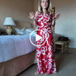 Fleur De Force stands in a hotel room in California. She wears a red and white cowl neck dress. Fleur appears in front of two large white and beige beds with her blonde hair loose and naturally curled.