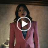 Colby Minifie stands in a stone wall room with black lamps and a dark wooden ceiling. She wears a structured pink blazer with a teal satin top.